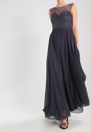 Robe de soirée bleu marine sans manches avec corsage orné de perles, encolure en tulle transparent, jupe fluide en chiffon, portée avec des talons noirs à brides.