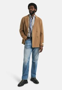 Beiger, doppelt gestreifter Wollmantel über einem mehrfarbigen gestreiften Hemd, kombiniert mit hellblauen Jeans und schwarzen Schnürschuhen.