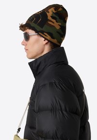 Piumino nero con colletto alto, abbinato a un berretto lavorato a maglia camouflage. Il modello indossa occhiali da sole scuri, mettendo in risalto uno stile urbano e raffinato.