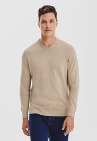 Beżowy sweter z dekoltem w kształcie litery V, wykonany z miękkiej dzianiny, z długimi rękawami i ściągaczem na dole. Nosi się go na ciemnoniebieskie dżinsy.
