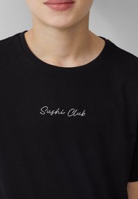 Zwart katoenen T-shirt met een ronde hals en korte mouwen, met "Sushi Club" in witte cursieve letters geborduurd op de borst.