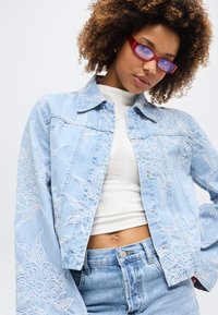 Νεαρή γυναίκα με σγουρά μαλλιά, που φοράει γυαλιά με κόκκινο φακό, ελαφρύ τζιν μπουφάν με κεντήματα, λευκό κοντομάνικο τοπ και τζιν παντελόνι που ταιριάζει.