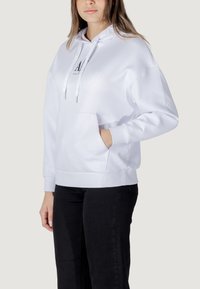 Sudadera blanca con capucha hecha de tela suave, con un bolsillo frontal, cordones ajustables y un logotipo impreso en el pecho.