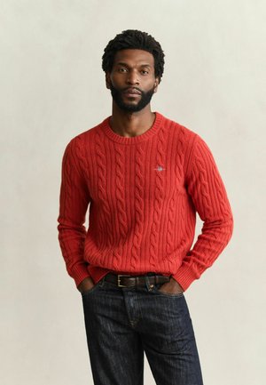 Homme barbu portant un pull rouge à torsades et un jean foncé, debout les mains dans les poches sur un fond uni.
