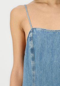 Lyseblå denimkjole med tynne stropper, rett utringning og avslappet passform. Stoffet har en falmet tekstur med synlige sømdetaljer.