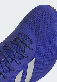Blue athletic shoe with a breathable fabric upper, featuring a textured pattern, flat laces, and silver accent stripes along the side.