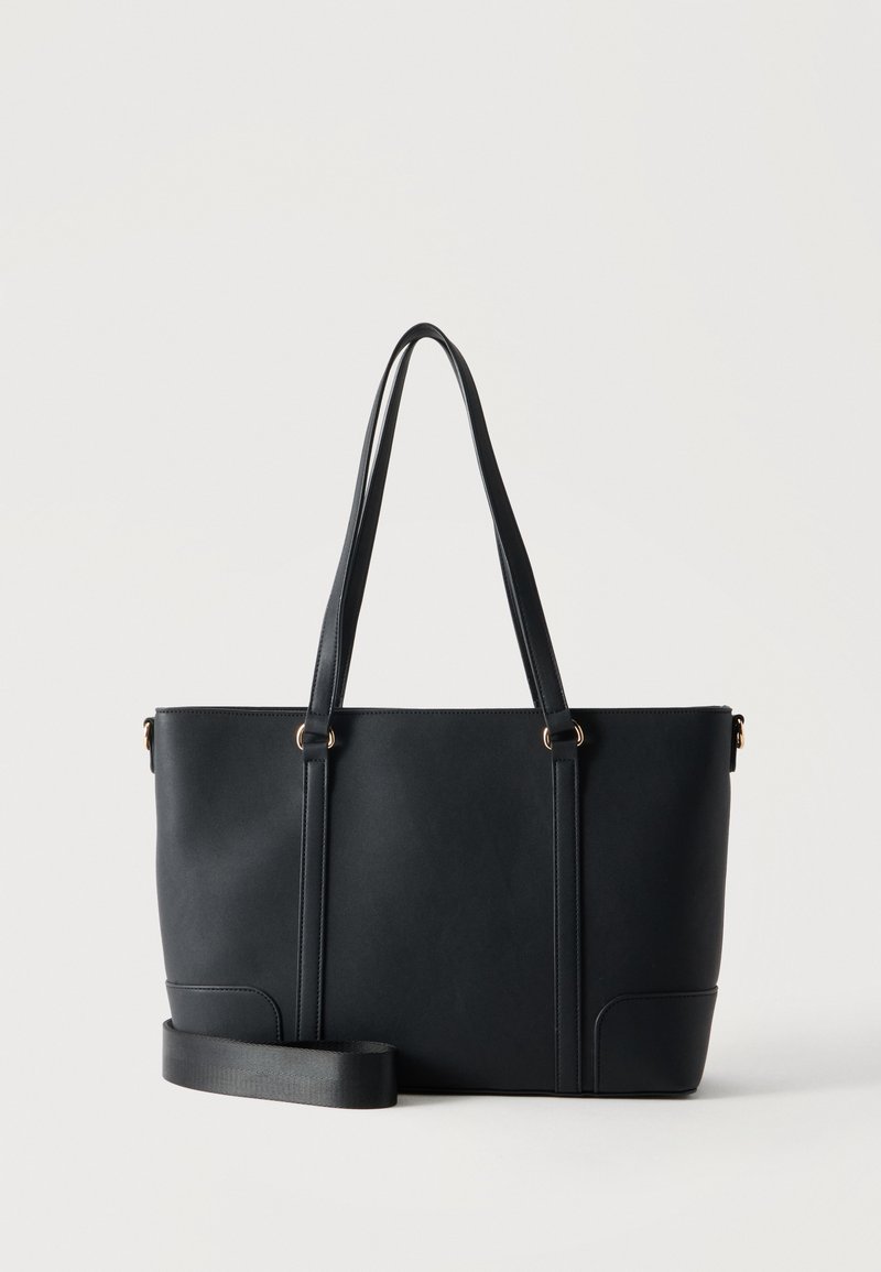 Sac cabas noir avec longues anses et une sangle d'épaule supplémentaire amovible sur un fond clair uni.