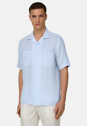 Jeune homme portant une chemise bleu clair à manches courtes avec deux poches poitrine et un pantalon blanc sur un fond uni.