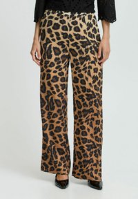 Pantalons larges à imprimé léopard en tissu beige et noir, offrant une texture lisse et un ajustement ample, associés à des chaussures noires.
