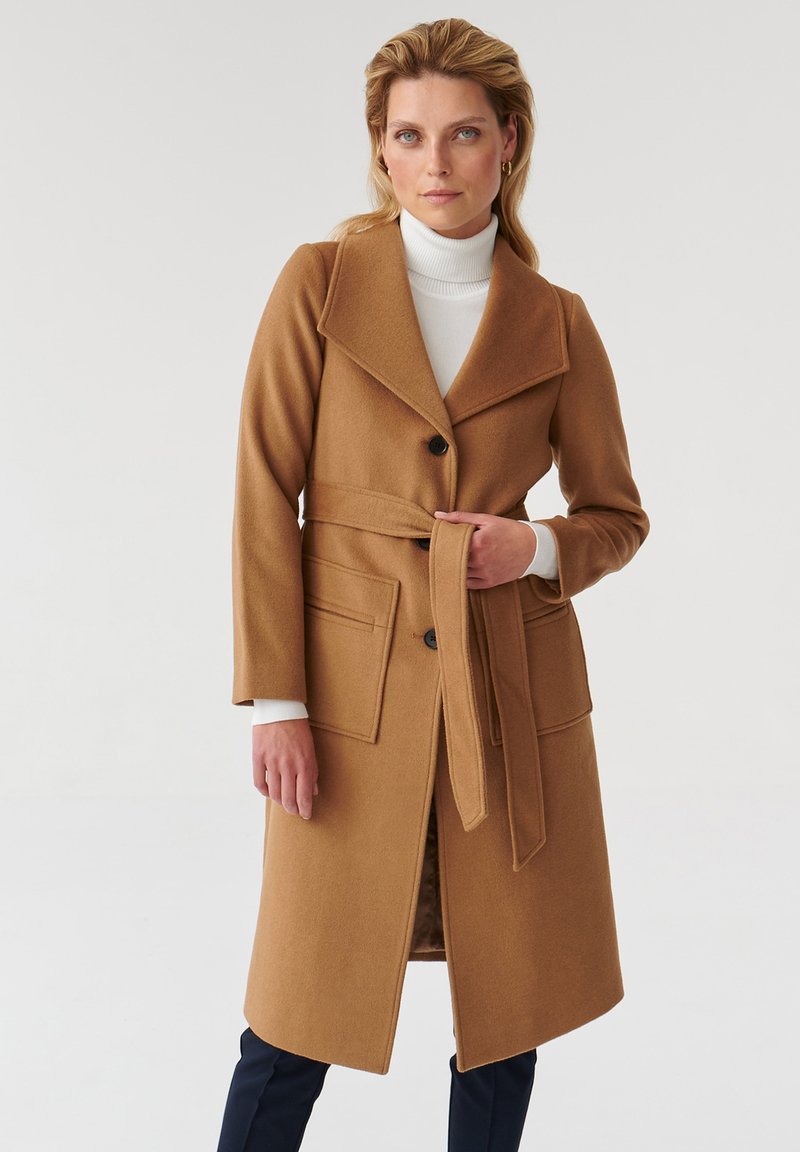 Cappotto di lana color cammello con ampi colli, chiusura a bottoni, cintura in vita e tasche frontali. Tessuto liscio e design sartoriale.