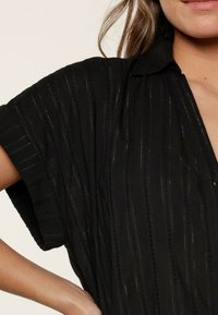 Maglietta nera a maniche corte con strisce verticali, colletto e sottili accenti testurizzati. Il tessuto sembra leggero con un design fluido.