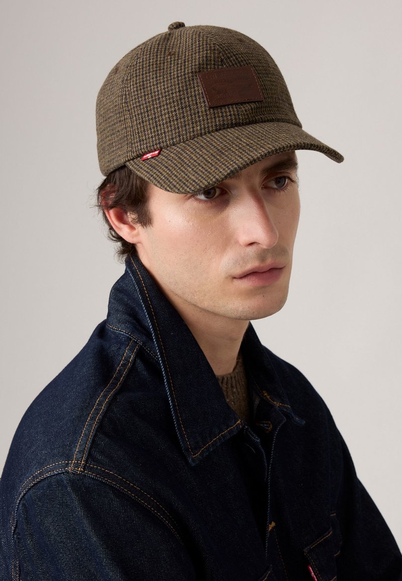 Brun rutigt keps med platt skärm, textil med struktur och en logotyp i brunt läder. Bärs med en mörk denimjacka med orange sömmar.