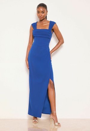 Full-length blue dress with a square neckline, cap sleeves, and a side slit. Smooth fabric with a fitted design. Model wears gold strappy heels.