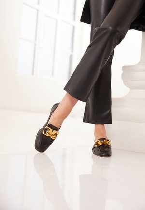 Beine in schwarzen Lederhosen und schwarzen Loafers mit großem goldenen Kettendetail, stehend auf einem reflektierenden weißen Boden.