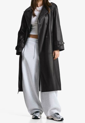 Personne portant un long manteau en cuir noir par-dessus un crop top blanc, un pantalon de survêtement large gris clair et des chaussures de sport argentées.