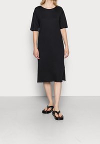 Svart kortärmad klänning med en lös passform, knälång, med en sidoklyvning. Bärs med svarta platta sandaler. Slät, lättviktig tyg.