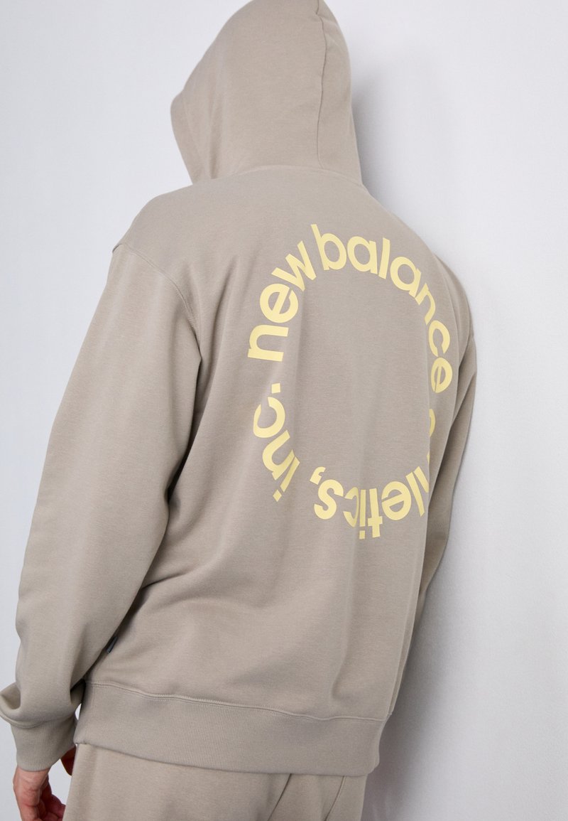 Hoodie marron avec un grand logo circulaire jaune au dos portant l'inscription "new balance". Fabriqué en coton doux et texturé.