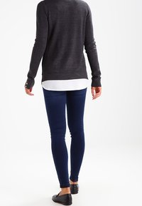 Tummanharmaa neulepusero, jossa on korkea kaulus, yhdistettynä tummansinisiin kapeisiin farkkuihin. Mustat litteät kengät ja valkoisen paidan helma näkyy alta.