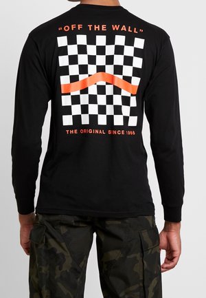 Mand iført sort langærmet skjorte med hvidt og orange ternet mønster og teksten "OFF THE WALL" og "THE ORIGINAL SINCE 1966" på ryggen.