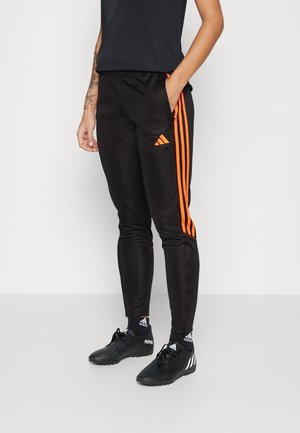 Persona con pantalones deportivos negros con rayas naranjas y logo de Adidas, calcetines negros de Adidas y zapatos negros con tacos, de pie con una mano en el bolsillo.
