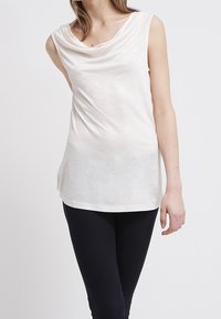 Top blanco sin mangas con escote drapeado, confeccionado en tela ligera, con una textura suave y un corte relajado, combinado con pantalones oscuros.