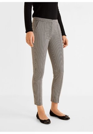 Frau trägt schwarze und weiße, schmal geschnittene Hahnentritt-Wadenhosen, kombiniert mit einem schwarzen Langarmoberteil und schwarzen Ballerinas mit Schleifen.