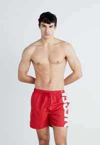 Shorts de bain rouges avec taille élastique à cordon de serrage, ornés d'un grand logo blanc sur le côté gauche. Tissu léger et longueur mi-cuisse.