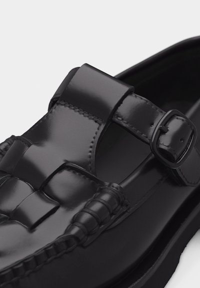 Sandale en cuir noir poli avec un détail tissé à l'avant et une bride réglable à boucle sur une semelle noire.