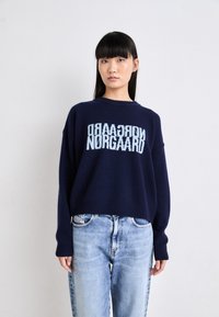 Maglione a maglia blu navy con bordo e polsini a coste, con la scritta "Nørgaard" in azzurro chiaro sulla parte anteriore. Vestibilità casual. Jeans in denim sotto.