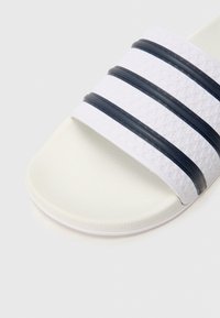 Λευκό σανδάλι με λευκή σόλα και τρεις μαύρες οριζόντιες ρίγες, υφή στην επάνω επιφάνεια και λεία σόλα. Το υλικό φαίνεται να είναι πλαστικό ή καουτσούκ.