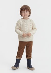 Junges Kind steht, trägt cremefarbenen, grob gestrickten Pullover, braune Cordhosen und dunkelblaue Gummistiefel mit beigen Sohlen.