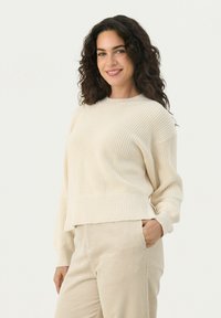 Gebreide crèmekleurige trui met ribbeltextuur, ronde halslijn en verlaagde schouders, gecombineerd met lichtbeige corduroy broeken.