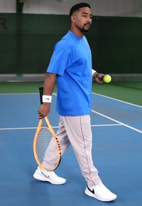 Tennis spiller i en blå skjorte og grå bukser holder et oransje racket og en grønn ball, iført hvite sko med svarte detaljer. Innendørs bane bakgrunn.
