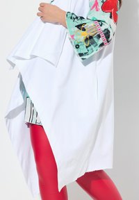 Maglietta bianca oversize con maniche decorate da motivi floreali in teal, rosso e giallo. Abbinata a leggings rossi lucidi. Taglio lungo e asimmetrico.