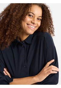 Lächelnde Frau mit lockigem Haar trägt ein marineblaues Hemd mit Knöpfen, die Arme verschränkt, vor einem schlichten weißen Hintergrund.