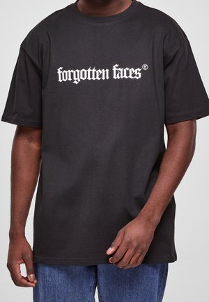 T-shirt noir à manches courtes avec texte blanc « forgotten faces » porté par une personne avec une main près de la taille, associé à un jean bleu.