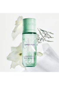 Bouteille en verre transparent avec un capuchon vert menthe. L'étiquette indique "CK Cotton Musk" en violet. Gouttes d'eau visibles et accents floraux en arrière-plan.