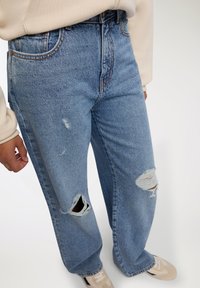 Jeans in denim chiaro con una vestibilità rilassata, caratterizzati da ampie strappi sulle ginocchia e un leggero sbiadimento. Abbinati a un sweatshirt beige e scarpe nei toni neutri.