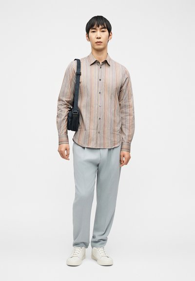 Άνδρας που φοράει ριγέ μακρυμάνικη μπλούζα, ανοιχτό γκρι παντελόνι, λευκά αθλητικά παπούτσια, κρατώντας μαύρη μικρή τσάντα ώμου σε απλό λευκό φόντο.