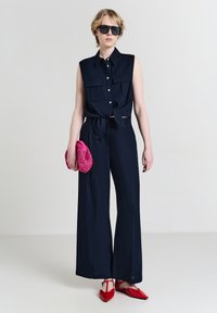 Persona con top senza maniche nero a bottoni e pantaloni a gamba larga, che tiene una borsa a mano rosa e indossa scarpe rosse con la punta e occhiali da sole neri.