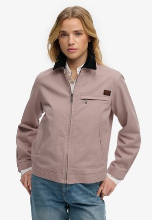 Kvinde med blondt hår iført lys mauve lynlåstrøje med sort krave og blå jeans, stående med hænderne på hoften.