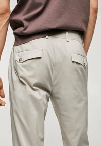 Un pantalon beige clair avec une texture lisse présente deux poches arrière, chacune avec une fermeture à bouton et un subtil détail de couture.