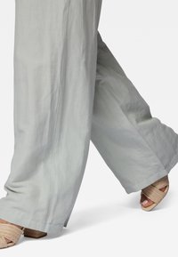 Lichtgrijze, wijde broek van een zachte linnen textuur, met een nette zoom en gedragen met geweven beige sandalen.