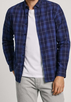 Mand iført en åben blåternet skjorte over en hvid t-shirt med lysegrå bukser, stående med den ene hånd synlig ved siden.