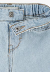 Lysblå denim med en samlet talje og metaltrykknaplukning. Viser kontrastsyninger og tekstureret stof i en nærbillede.