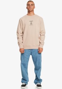 Sweatshirt beige clair avec des poignets et un ourlet côtelés, orné d'un motif graphique sur la poitrine, associé à un jean bleu clair à coupe décontractée et des baskets noires.
