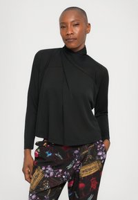 Zwarte lange mouw top met een opgevouwen kraag en een zachte textuur, gecombineerd met donkergekleurde broek met een patroon en bloemdecoraties in levendige kleuren.