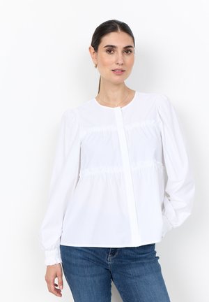 Frau mit dunklem Haar, die eine weiße langärmelige Bluse mit gerafften Details und blaue Jeans vor einem schlichten weißen Hintergrund trägt.