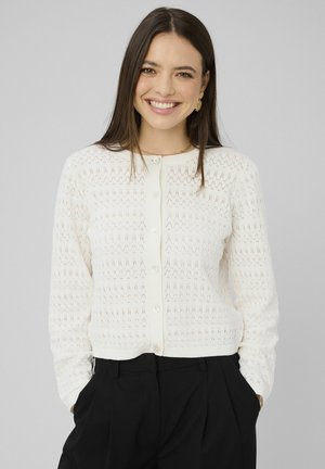 Jeune femme souriante, portant un cardigan en tricot blanc boutonné et un pantalon noir, les mains dans les poches, debout devant un fond uni gris clair.