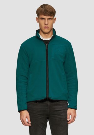 Jonge man in een teal fleece jack met rits en zijzakken, voorzien van een subtiele borstlogo, gecombineerd met zwarte jeans, staande tegen een effen achtergrond.